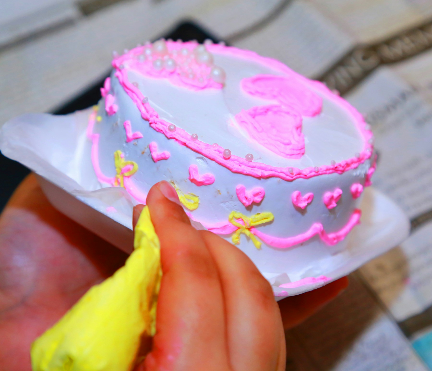 A hand holding a piping bag while decorating a small cake with pink icing