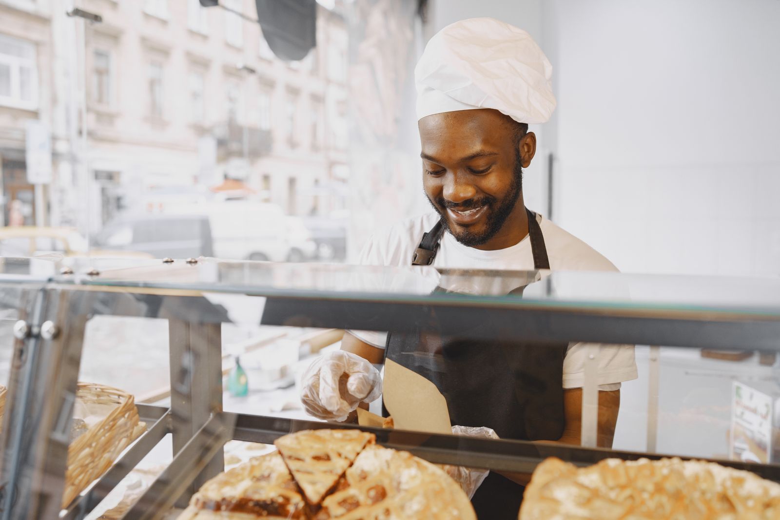 A baker wearing a white chef hat and gloves behind a glass pastry display.